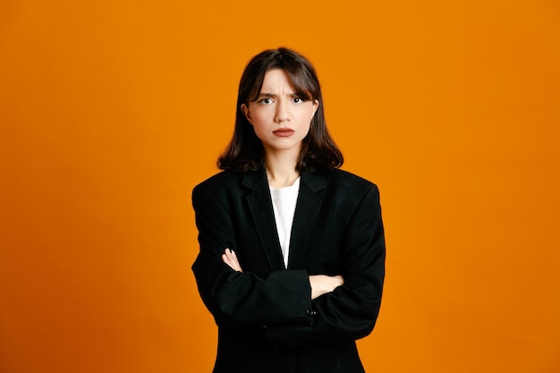 strict crossing hands young beautiful female wearing black jacket isolated on orange background