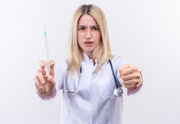 Strict doctor young blonde girl wearing stethoscope and medical gown holding syringe holding out fist to camera on isolated white background