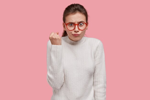 Strict European lady with pleasant look, shows fist , wears optical glasses and white jumper, demonstrates her dislike
