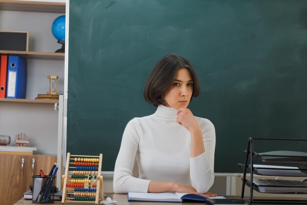 교실에서 학교 도구를 들고 책상에 앉아 있는 엄격한 턱 젊은 여교사