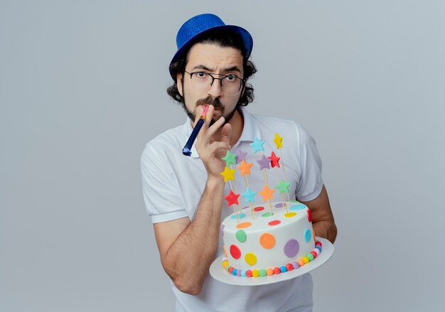 Strict handsome man wearing glasses and blue hat holding cake and blowing whistle isolated on white