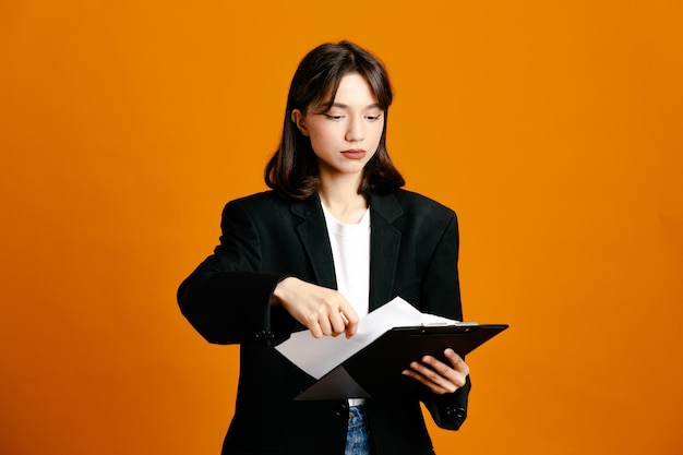 strict holding clipboard young beautiful female wearing black jacket isolated on orange background