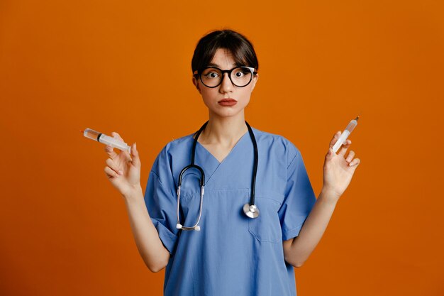 オレンジ色の背景で隔離の均一なフィス聴診器を身に着けている厳密な保持注射器若い女性医師