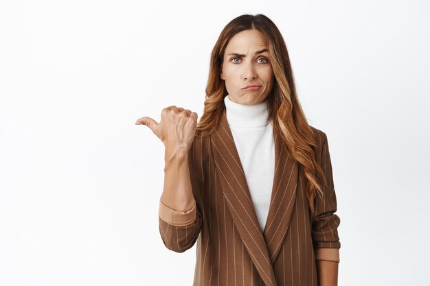 Strict lady boss looking suspicious pointing left and staring with skeptical face at camera standing in brown suit over white background