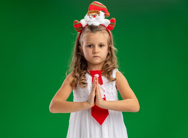 Строгая маленькая девочка в рождественском обруче для волос с галстуком, показывающим жест молитвы на зеленом фоне