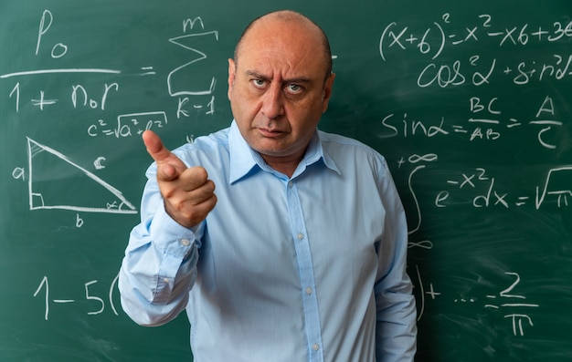 Strict looking at camera middle-aged male teacher standing in front blackboard showing you gesture