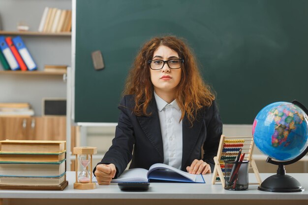 教室で学校の道具を持って机に座って眼鏡をかけている若い女性教師のカメラを厳しく見る