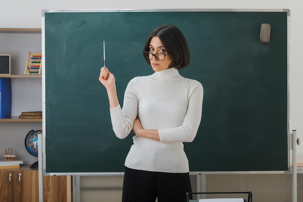 strict looking at camera young female teacher wearing glasses standing in front blackboard and points at blackboard with pointer in classroom