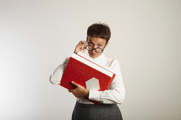 strict looking teacher in white blouse, gray tweed shirt and black round glasses holding bright red and white binders looks suspiciously above glasses and makes a skeptical face