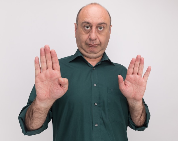 strict middle-aged man wearing green t-shirt showing stop gesture isolated on white wall