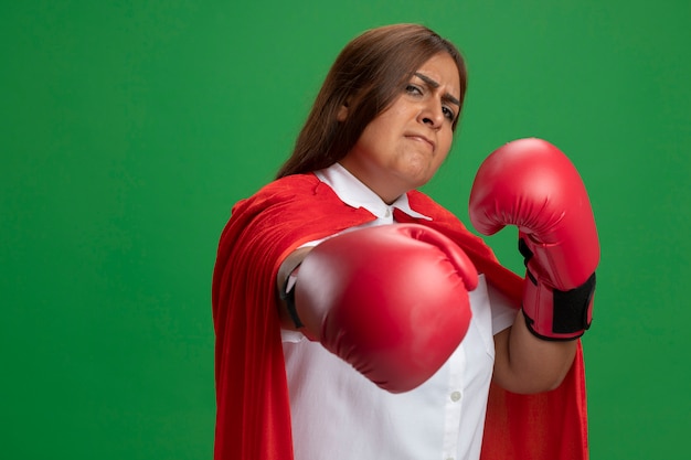 Strict middle-aged superhero female wearing boxing gloves holding out hand isolated on green