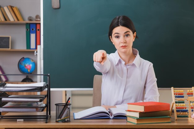 strict points at camera young female teacher sitting at desk with school tools in classroom