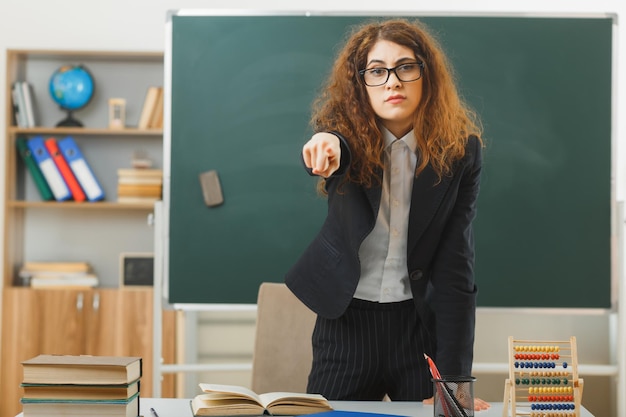 Foto gratuita punti severi alla telecamera giovane insegnante femminile in piedi davanti alla lavagna in classe