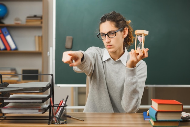 Foto gratuita punti severi a lato giovane insegnante maschio che tiene un orologio di sabbia seduto alla scrivania con gli strumenti della scuola in classe