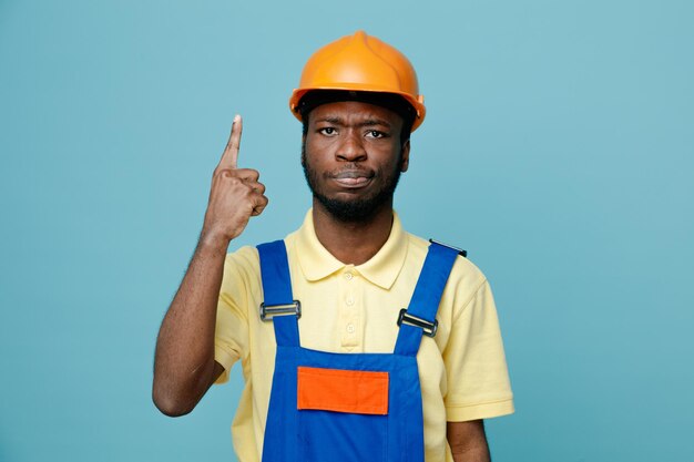 Strict points at up young african american builder in uniform isolated on blue background