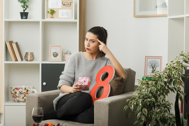 Strict putting finger on temple beautiful girl on happy women day holding present sitting on armchair in living room