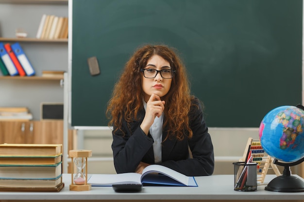 教室で学校の道具を使って机に座っている眼鏡をかけている若い女性教師のあごに厳密に手を置く