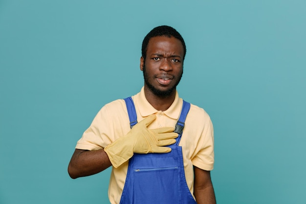 Strict putting hand on heart young africanamerican cleaner male in uniform with gloves isolated on blue background