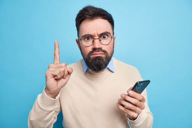 Strict serious bearded adult man raises index finger has excellent idea uses new mobile application holds smartphone wears spectacles and sweater 