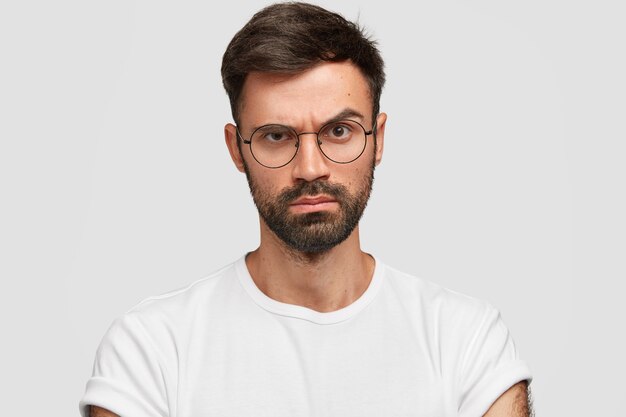 Strict serious discontent male boss raises eyebrows with angry displeased expression, dislikes something, expresses negative emotions, dressed casually, poses against white wall.