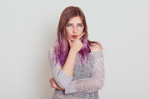 あごに指を入れた薄紫色の髪の厳格な真面目な女性、重要なことやトラブルについて考え、スタイリッシュなシャツを着て、灰色の壁に孤立してポーズをとる。