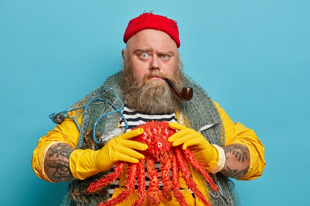 Strict serious man with thick beard, holds big red crab, smokes tobacco pipe, enjoys sailing and cruise, wears red hat, fishing net over shoulders