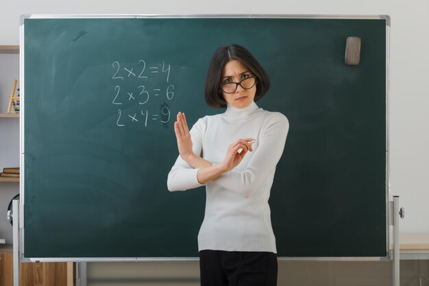 엄격한 제스처를 보여주는 젊은 여교사는 안경을 쓰고 칠판 앞에 서서 교실에서 글을 씁니다.