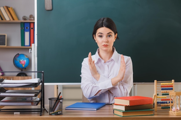 strict showing no gesture young female teacher sitting at desk with school tools in classroom