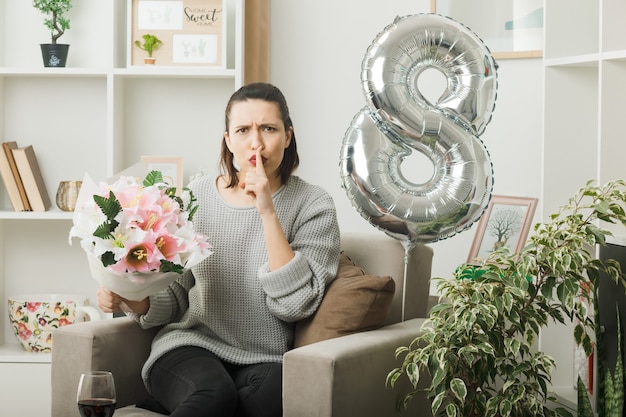 Strict showing silence gesture beautiful girl on happy women day holding bouquet sitting on armchair in living room