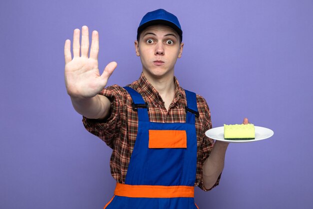 Strict showing stop gesture young cleaning guy wearing uniform and cap holding sponge on plate 