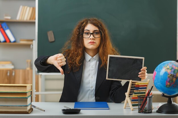 strict showing thumbs down young female teacher wearing glasses holding mini chalkboard sitting at desk with school tools in classroom