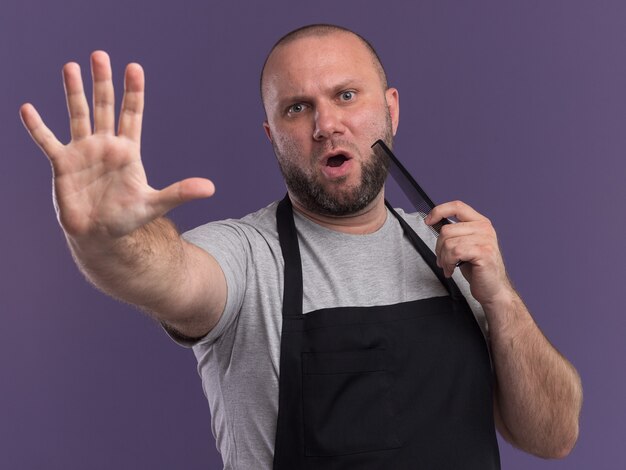 Strict slavic middle-aged male barber in uniform combing beard showing stop gesture isolated on purple wall