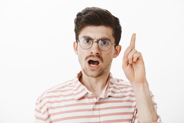 Strict teacher scolding students for bad behaviour. Shot of serious focused bearded male with moustache, raising index finger while giving speech and arguing, standing displeased over gray wall