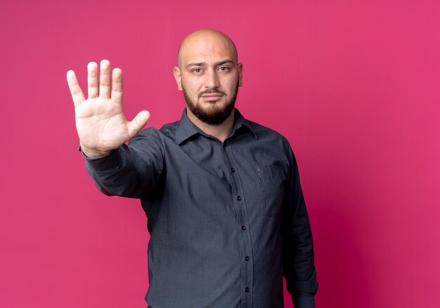 Strict young bald call center man doing stop gesture at front isolated on crimson wall