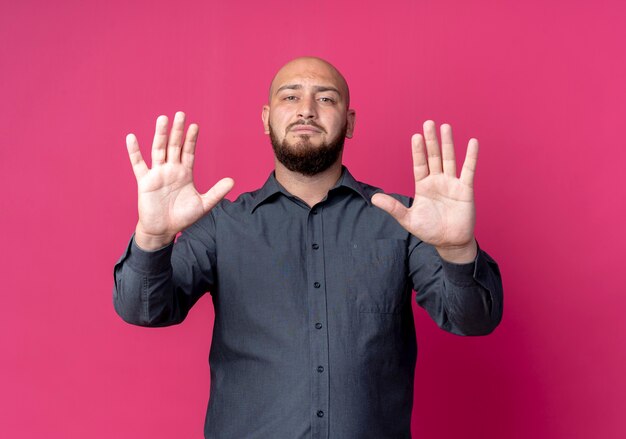Strict young bald call center man stretching out hands at front gesturing stop isolated on crimson wall