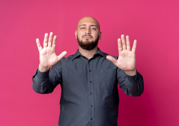 Strict young bald call center man stretching out hands at front gesturing stop isolated on crimson wall