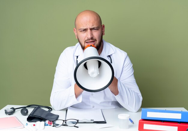 Strict young bald male doctor wearing medical robe and stethoscope sitting at desk work