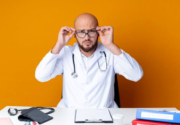 Strict young bald male doctor wearing medical robe and stethoscope sitting at work desk with medical tools take on glasses on orange