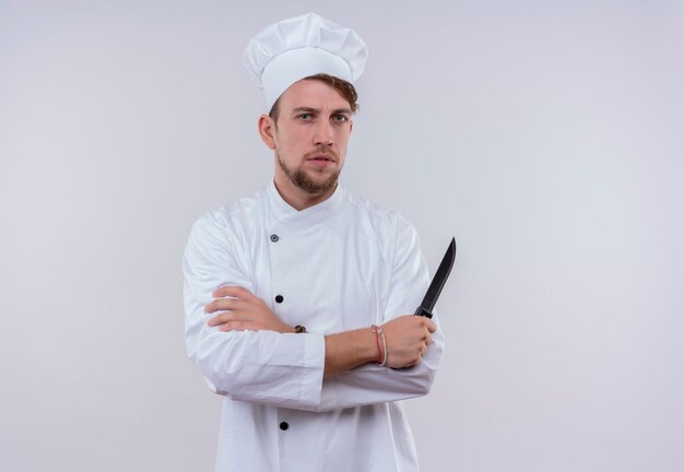 A strict young bearded chef man in white uniform holding a knife with folded hands while looking on a white wall