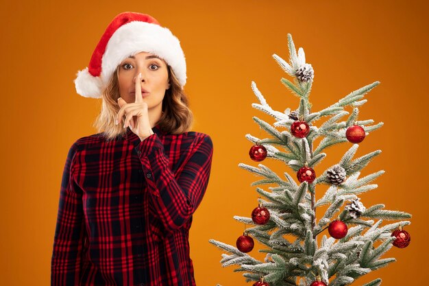 Strict young beautiful girl standing nearby christmas tree wearing christmas hat showing silence gesture isolated on orange background