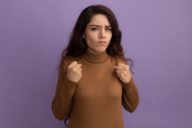 Strict young beautiful girl wearing brown turtleneck sweater holding fists isolated on purple wall