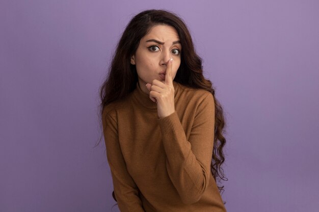 Strict young beautiful girl wearing brown turtleneck sweater showing silence gesture isolated on purple wall