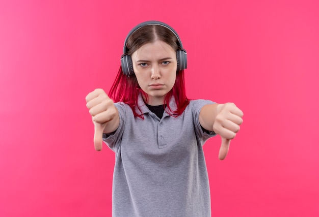 Strict young beautiful girl wearing gray t-shirt in headphones her thumbs down on isolated pink background