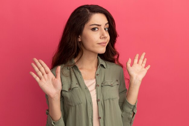 Strict young beautiful girl wearing olive green t-shirt spreading hands isolated on pink wall