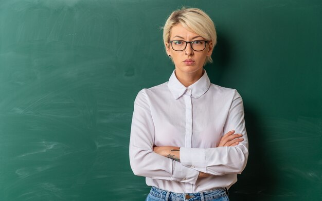 Strict young blonde female teacher wearing glasses in classroom standing with closed posture in front of chalkboard  with copy space