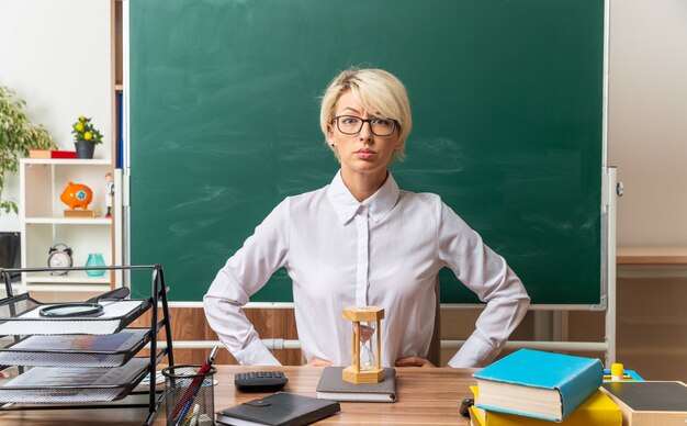 机に座って教室で学用品を持って眼鏡をかけている厳格な若いブロンドの女性教師は、正面を見て腰に手を保ちます