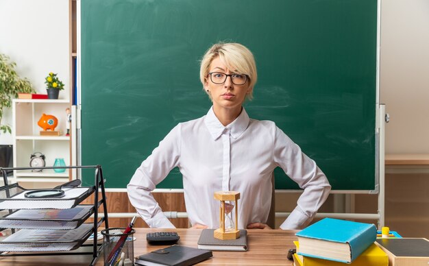 строгая молодая блондинка учительница в очках сидит за столом со школьными принадлежностями в классе, держа руки на талии, глядя вперед