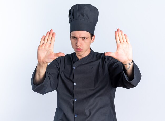 엄격한 젊은 금발 남성 요리사 유니폼과 모자 중지 제스처를하는 요리사