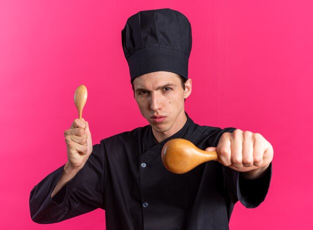 Strict young blonde male cook in chef uniform and cap holding and stretching out wooden spoons towards camera 