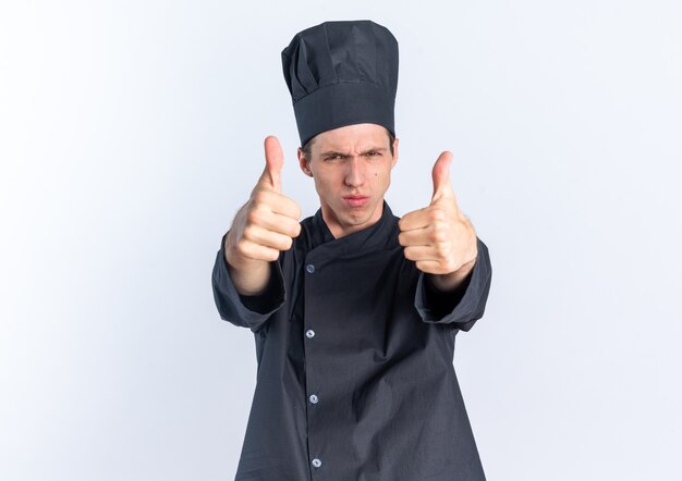 엄격한 젊은 금발 남성 요리사 제복을 입고 흰색 벽에 고립 된 엄지 손가락을 보여주는 카메라를 보고 모자